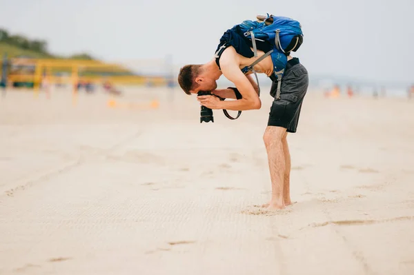 一个穿着短裤 背着背包 赤身裸体的小伙子在拍摄沙子的时候 靠在后面调整相机的镜头 摄影记者寻找好的相框 — 图库照片