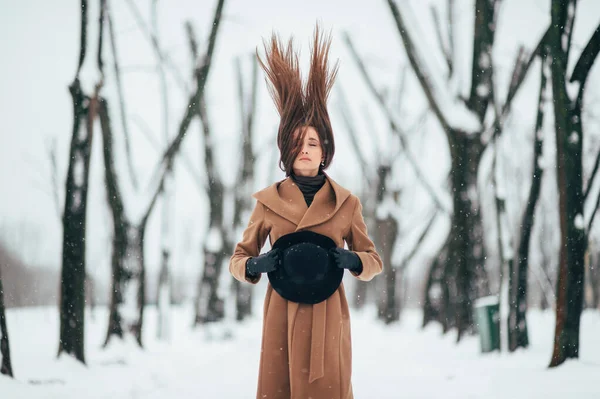 Unusual portrait of beautiful girl with dramatic face holding her hat and trowing up her hair in winter tree valley