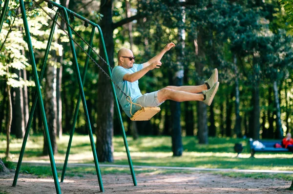 滑稽的成年秃头男子戴着太阳镜 从地面上摆动时跳了起来 童年的行为夏日 在城市公园的户外儿童游乐场里 一个笨笨可笑的大男孩儿在玩耍 — 图库照片