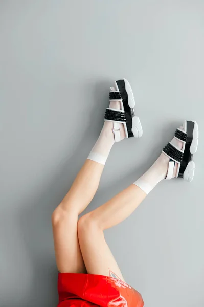 Beautiful female legs in white mesh trendy socks wearing fashionable black white high wedge leather sandals. Womens modern fashion footwear. Rebel girl in red leather skirt on gray background
