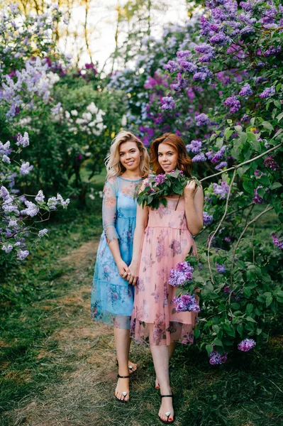 ロマンチックな気分だ 双子の姉妹は優しい関係 ライラックガーデンでカメラのポーズをとるカラフルな夏のドレスの美しい双子の若いモデル 陽気なかわいい妖精の女の子でマジックパーク屋外 — ストック写真