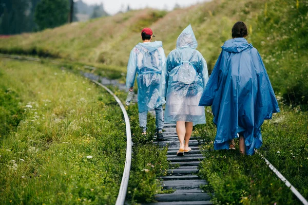 Crew of informal young travelers in raincoats walking in danger zone on railroad outdoor at nature. Company of friends rest on vacation. People go on railway sleepers. Cloudy muggy weather