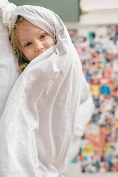 Vetrtically turned portrait of little cheerful girl with funny face  lying on bed at home under white blanket and looking ar camera with decorative wall on background. 