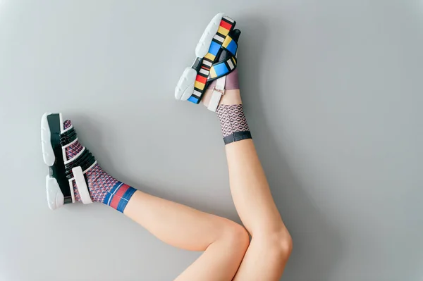 Beautiful female legs in mismatched trendy socks posing in two different fashionable high wedge leather sandals on gray background. Odd disargonized young girl wearing high sole summer stylish shoes