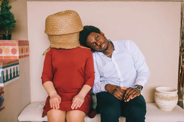 Bizarre interracial loving couple. Dark skinned african man with smiling face sitting with his caucasian unrecognizable girlfriend with straw basket on her head at home. Weird intternational relationship.