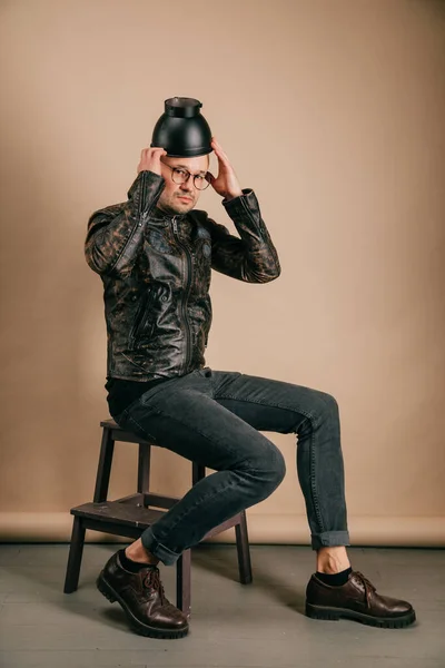 茶色の背景にスタジオで頭の上に黒い鍋で木製の椅子に座ってファッショナブルな服の若いスタイリッシュな男の面白い肖像画 革のジャケット ブローグシューズでハンサムな笑顔の男の子 — ストック写真