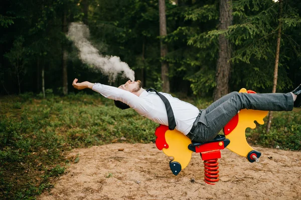 Adult serious man in business clothes riding childrens metal horse attraction with spring on playground. Odd person blows out clouds of thick smoke. Strange posing with arms apart. Weird behavior.