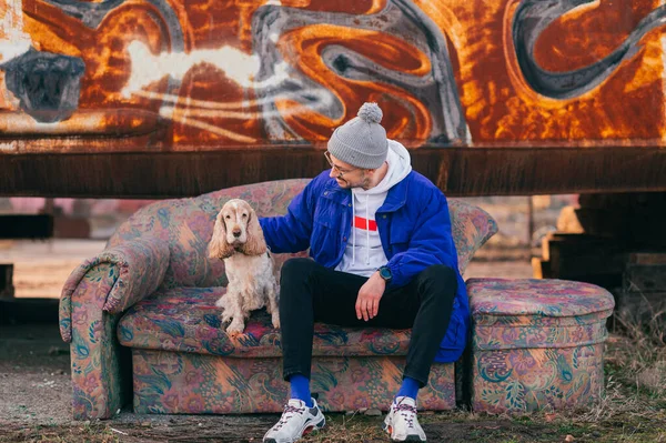 Stylish young man in blue old fashioned jacket, white hoodie and modern sneakers siiting on old couch with best friend pet - cocker spaniel dog over rusty metal graffiti wall on background.