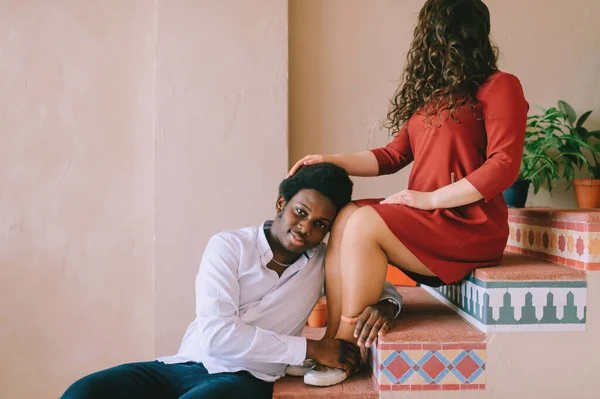 Weird interracial couple. Happy adult smiling dark skinned man siiting on stairs in decorative room and hugging legs of his caucasian girlfrined with face covered by hair. Portrait of strange lovers