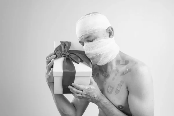 Injured man with many patches on his body and bandaged head sits and holds a white box with gift isolated on white background