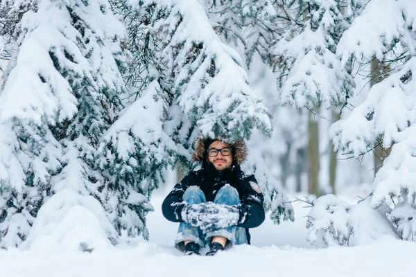 大人の男は森の屋外で木の下に雪の中に座っている 冬の髭の男は1月に自然の中でリラックスしたパーカーをフード付き カメラを見ながら眼鏡をかけて笑顔で前向きな少年 凍えてる人 — ストック写真