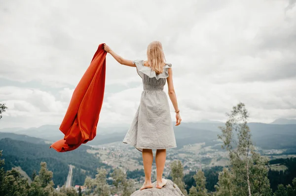 Woman in wild mountains gives distress signal SOS using red cover. Concept of emergency situation during hike in mountains. Barefoot woman stands at stone, waving red blanket and waiting for help.