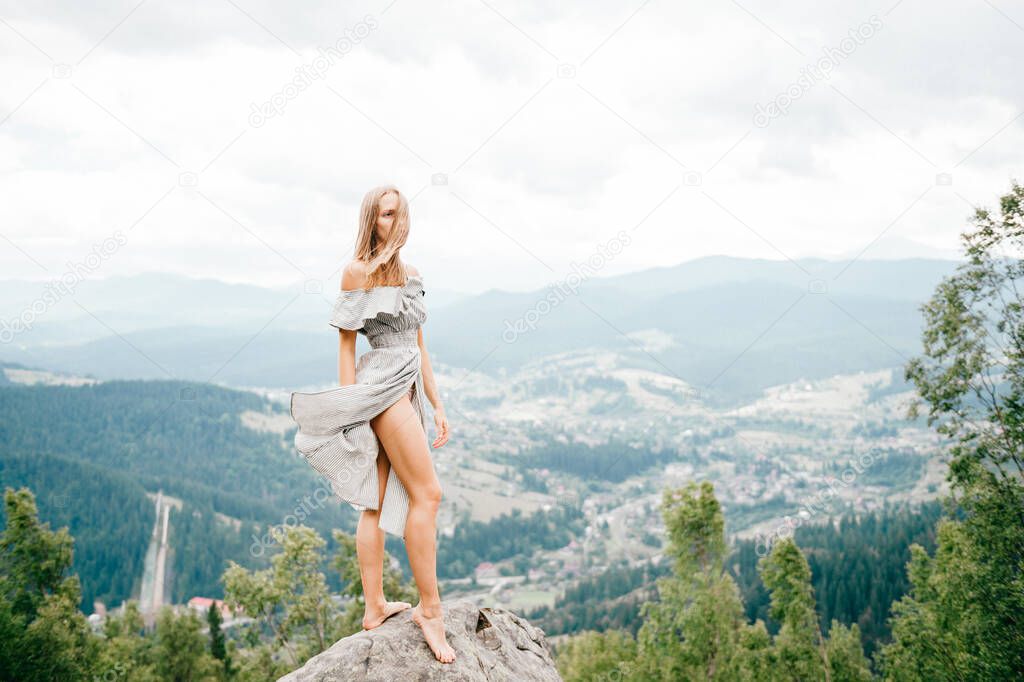 Young beautiful barefoot blonde girl with long hair in summer dress standing on top of conquered mountain at stone and enjoying fabulous landscape scenic view with mountains and village in valley 