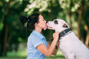 Uzun siyah saçlı, mavi elbiseli güzel yüzlü beyaz bir kızın portresi güzel siyah beyaz köpeğiyle parkta oturuyor ve gülümsüyor.