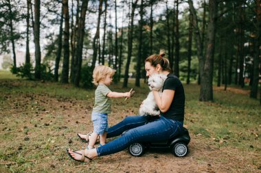 Genç anne, büyük yaz ormanında küçük çocuğu ve beyaz köpeğiyle oynuyor..