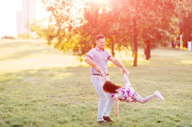 Babam kızımsı elini bacağıyla tutuyordu ve dönüyordu. Heyecanlı aile yaz parkında iyi eğlenceler. Babam yeşil çayırda küçük kız çocuğuyla oynuyor. Kaygısız mutlu bir çocukluk. Babalık 