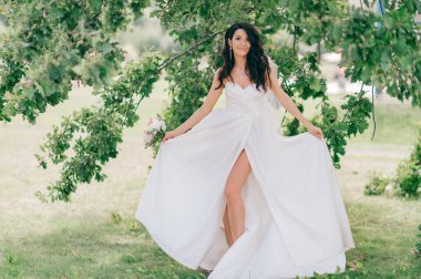 Güzel beyaz elbisesiyle mutlu bronzlaşmış gelin yaz günü bir buket çiçekle ağacın önünde poz veriyor.