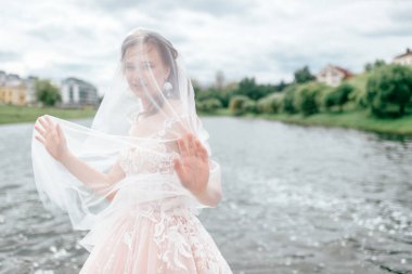 Yüzü peçeyle örtülmüş, arka planda gölle dışarıda poz veren gelinlik giymiş güzel bir gelin..