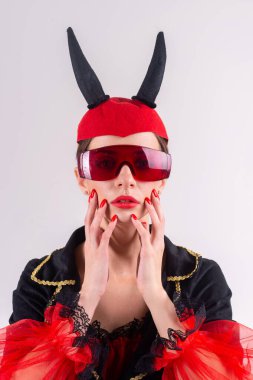 Fütürist gözlüklü, siyah kırmızı karnaval kostümlü ve kafasında şeytan boynuzları olan cazibeli seksi kadının stüdyo moda portresi. 