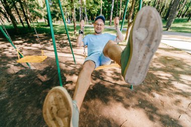 Garip, tuhaf, tuhaf insanlar konsepti. Gözlüklü, komik duygusal yüzlü, yetişkin stil sahibi bir adam yazın çocuklar için oyun parkında salıncakta sallanıyor. Mutlu insan çocukluğun tadını çıkarıyor