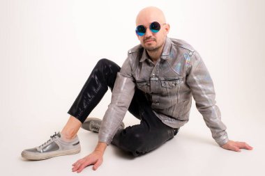 Beyaz saçlı, gri gömlekli, siyah deri pantolonlu ve siyah gözlüklü gri spor ayakkabılı şık beyaz bir adam yerde oturuyor ve beyaz arka planda izole edilmiş kamera için poz veriyor..