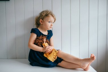 Trubschachen, İsviçre - 16 Aralık 2019: Mavi elbiseli, kısa saçlı güzel beyaz kız sevimli barbekü oyuncağını tutuyor, parlak bebek odasında oturuyor ve gülümsüyor.