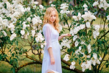 Beyaz leylak ağacının yanında uzun mavi elbiseli genç beyaz kadın duruyor.