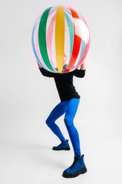 Spor kıyafetleri içinde şık bir adam, plaj topunu suratının önünde beyaz stüdyo arka planında tutuyor..