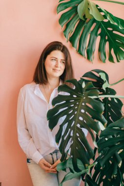 Güzel esmer kadın, dinleniyor ve pembe duvarda kameraya poz veriyor. Mutlu, mutlu, gülümseyen beyaz kadın portresi yeşil dalları olan palmiye ağacının yanında duruyor.