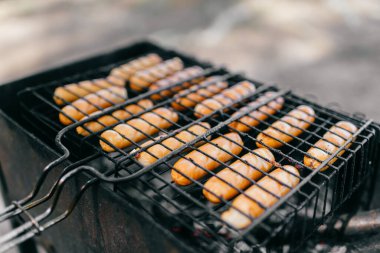 Sosisler ızgarada, kömürün üstünde, dumanla kaplanmış, açık havada. Lezzetli çerez orman gezgini. Güneşli bir günde kömürlü sosisli piknik.