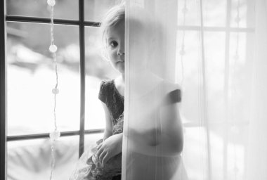 Beyaz, siyah, beyaz, sarı saçlı, mavi elbiseli küçük bir kızın güzel oyuncağını tutuyor ve bebek odasında pencere kenarında oturuyor ve gülümsüyor..