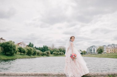 Yüzü peçeyle örtülmüş, arka planda gölle dışarıda poz veren gelinlik giymiş güzel bir gelin..