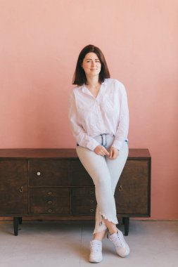 Güzel esmer kadın, dinleniyor ve pembe duvarda kameraya poz veriyor. Evdeki ahşap klozette oturan neşeli gülümseyen mutlu beyaz kadın portresi.