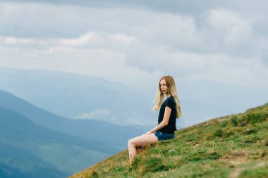 Genç kız dağın tepesinde rahatlıyor ve manzaranın tadını çıkarıyor. Mutluluk, mutluluk. Güzel yaz doğa manzarası. Yalnız sarışın genç gezgin tatilde dışarıda rüya görüyor. Romantik ruh hali