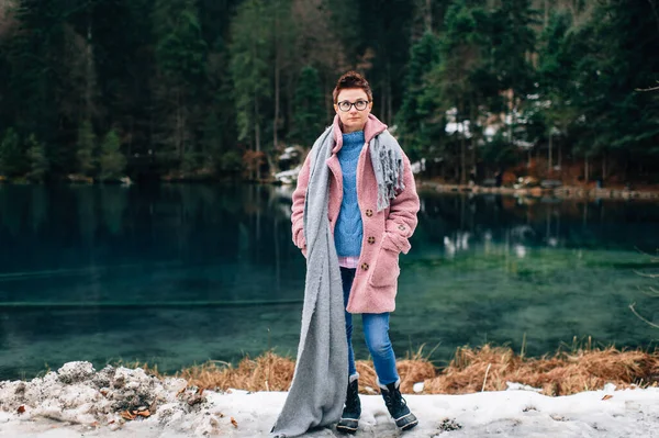 Picture of handsome woman in warn pink coat, denim trousers and with long grey scarf stands near the beautiful lake and smiles