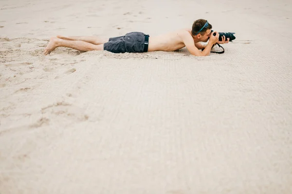 休暇で海の近くのビーチに横たわっている若い旅行者の男と自然の写真を撮る カメラと遠くから人々をスパイ大きなレンズを持つトップレスハンサムな裸足の男 趣味の時間だ 砂の上に眠る少年 — ストック写真
