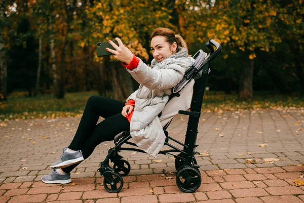 Funny picture of a young mom riding a pram in the park in the fall and taking lots of selfies on her new phone.