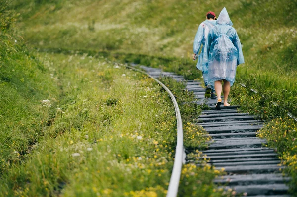 两个穿着雨衣的年轻旅行者走在长满青草的铁路上 一群朋友在度假时休息 旅游服装 多云的闷热天气 希波斯特的生活方式从角度来看哭 — 图库照片