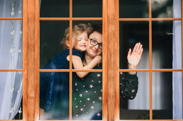 Portrait of beautiful young caucasian female with pretty face, short dark hair, big eyes, glasses with her cheerful child and looks through big window