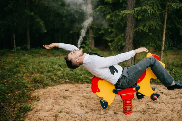 Adult serious man in business clothes riding childrens metal horse attraction with spring on playground. Odd person blows out clouds of thick smoke. Strange posing with arms apart. Weird behavior.