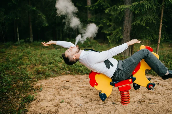 Adult serious man in business clothes riding childrens metal horse attraction with spring on playground. Odd person blows out clouds of thick smoke. Strange posing with arms apart. Weird behavior.