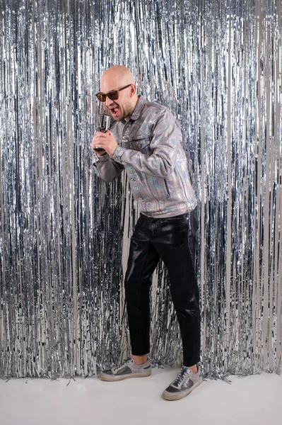 Beautifull caucasian man without hair with black sunglasses in a shiny shirt, black leather pants and grey sneakers sings in a small silver microphone.