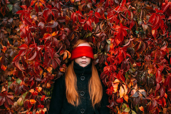 Portrait of longhaired redhead girl with red blindfold in her eyes standing against ivy wall in autumn park.