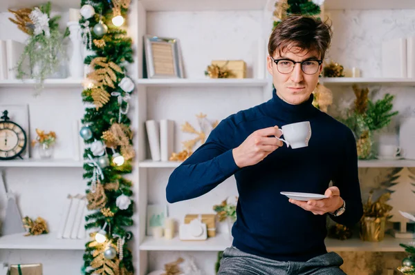 カメラのためのポジティブな感情的な顔を持つエレガントな男クリスマスツリーと新年の装飾と居心地の良い部屋でコーヒーを飲みながらポーズ — ストック写真