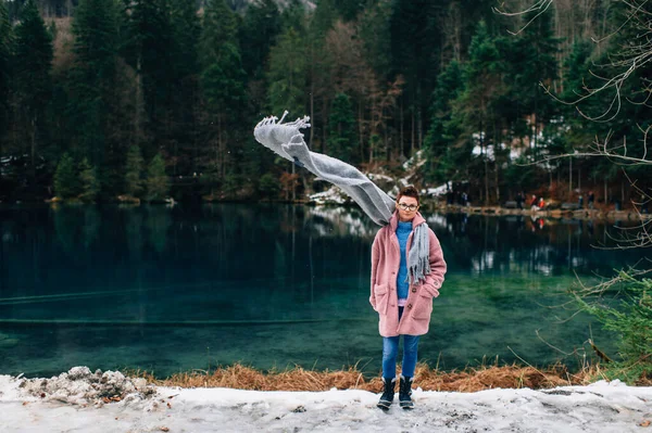 Picture of handsome woman in warn pink coat, denim trousers and with long grey scarf fluttering in the wind stands near the beautiful lake and smiles