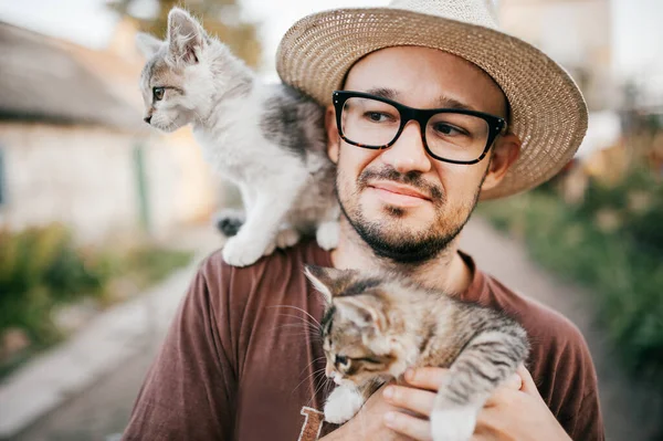 抽象的な背景を持つ村の屋外で手に2匹の小さな子猫を保持する幸せな若いひげを生やした農家 ガラスの笑顔の男と面白いかわいいペットと遊んでわら帽子 田舎でお楽しみください — ストック写真