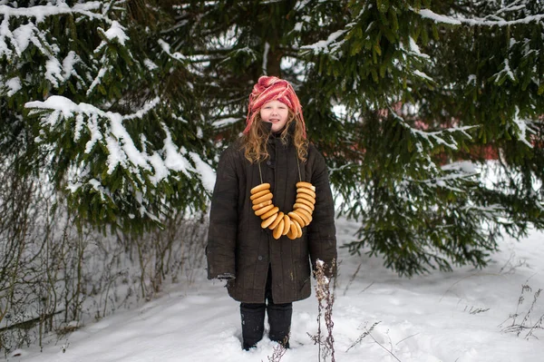 可愛い白人の村の女の子は冬に乾燥したリング状のロールサンドの笑顔で雪の森の中に立っています — ストック写真