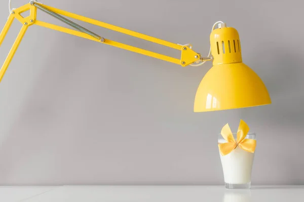 Glass of milk decorated with yellow bowtie standing on table under yellow lamp.