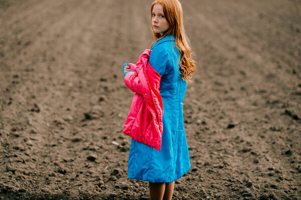 Beautiful strange red head girl in long blue coat and pink warm jacket spends a lot of time on the big empty field