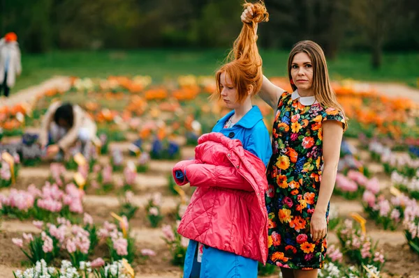 Pretty female child with red hair in blue coat stands in the big garden with her elder sister.